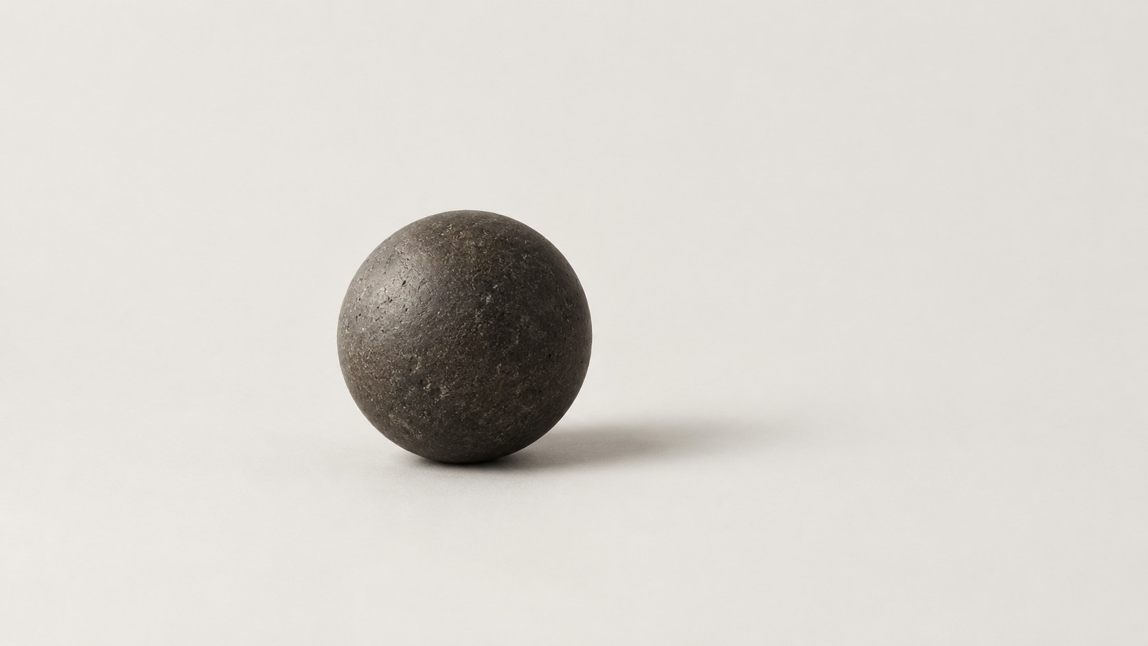 A single dark stone ball resting on a warm white ground, with a soft shadow beside it.