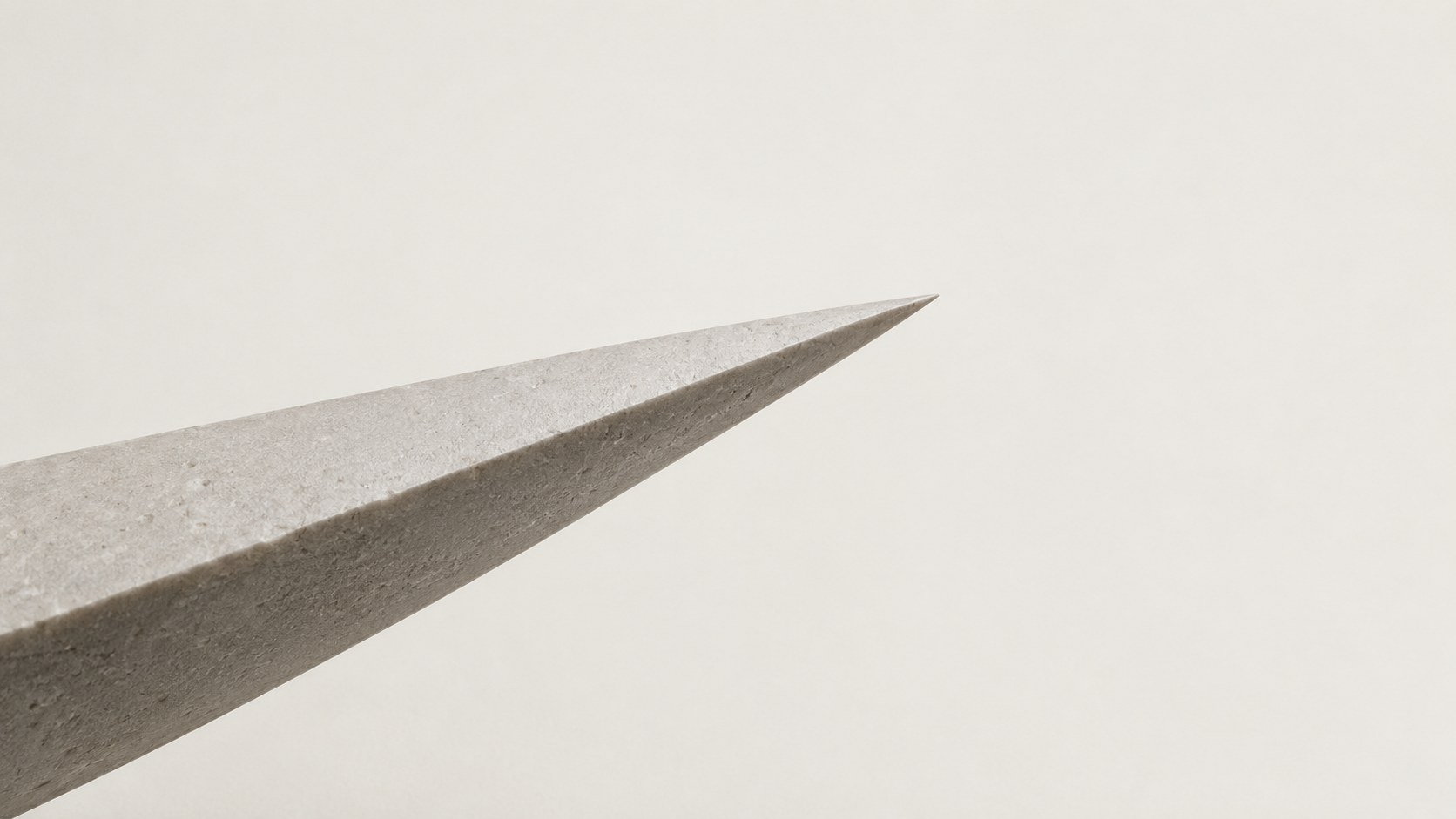 The sharp point of a pale stone blade against a warm white ground, its underside in soft shadow.