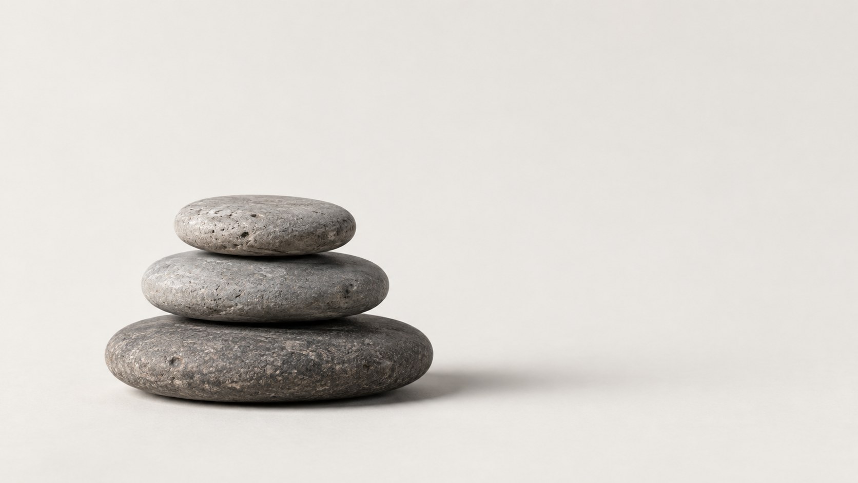 Three flat grey stones stacked in a small balanced pile on a warm white ground, with a soft shadow to the right.
