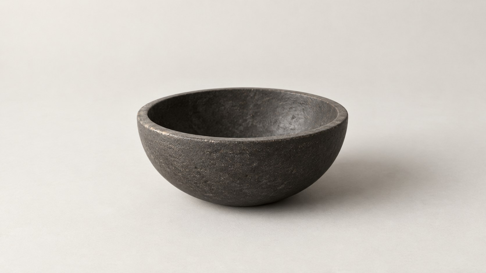 A shallow dark stone bowl on a warm white ground with a soft shadow to the right.