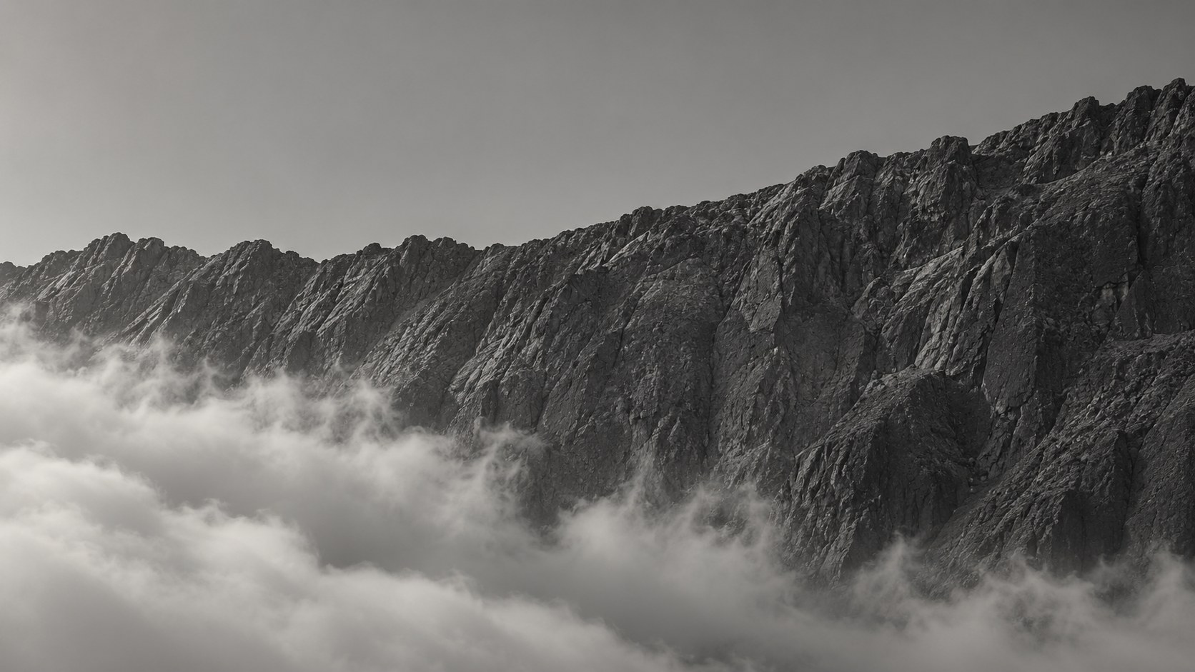 A long dark rock ridge with a jagged crest, rising out of pale cloud under an even grey sky.