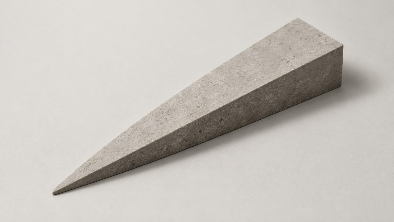 A pale stone wedge with a sharp point, lying at a slant across a warm white ground, with a soft shadow beneath it.