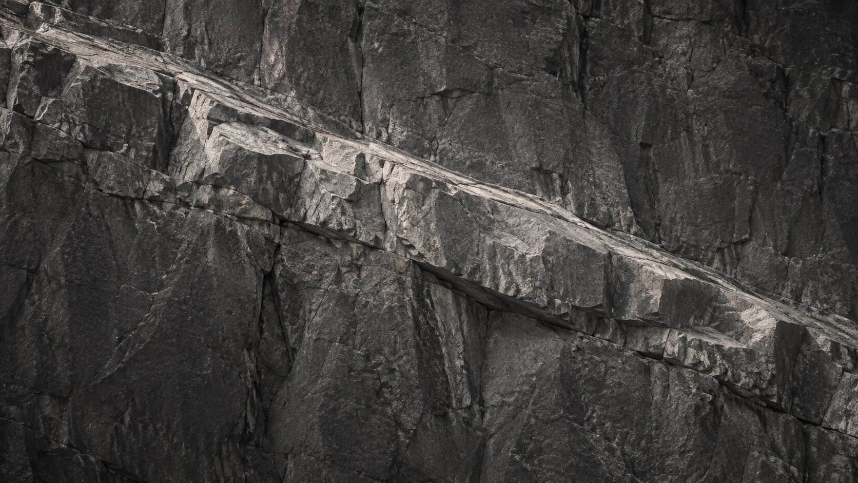 A steep dark grey rock face with a broad pale ledge crossing it, lit from the upper left.