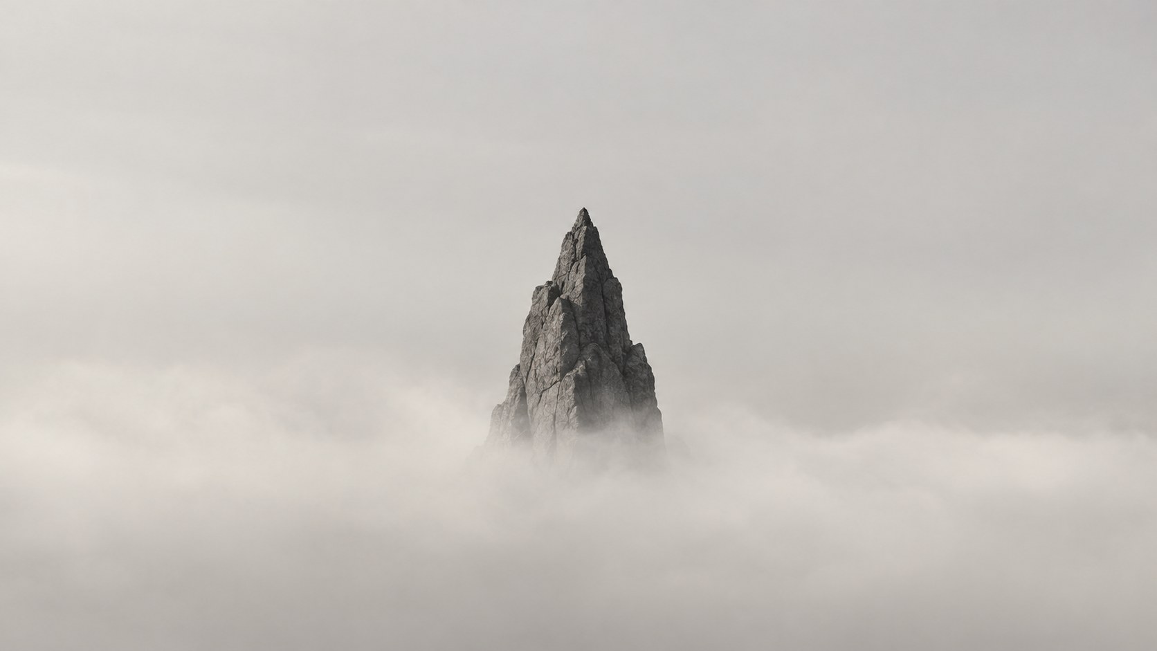 A small dark rock spire rising out of pale mist, under a plain pale grey sky.