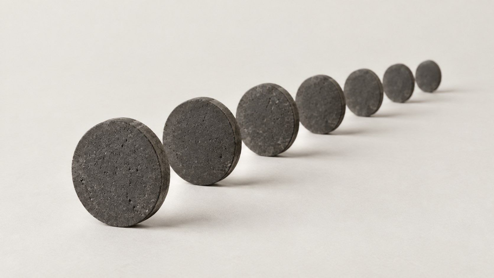 A row of identical flat dark stone discs standing on edge, getting smaller into the distance, on a warm white ground.