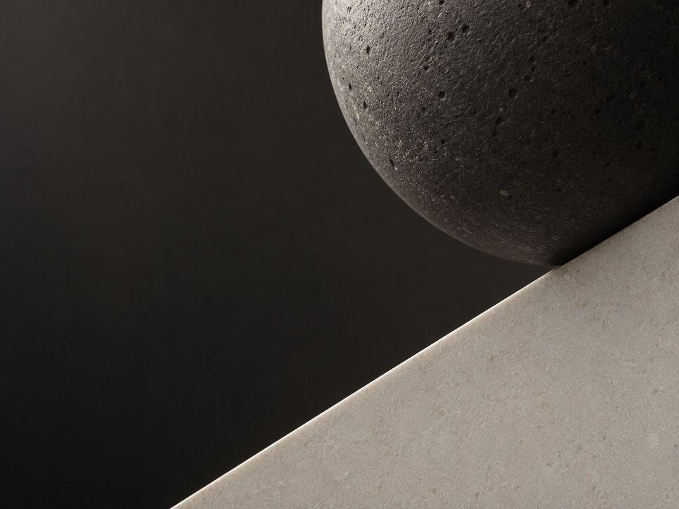 A close view of a dark stone ball touching the slanted edge of a thin pale stone blade, against a dark ground.